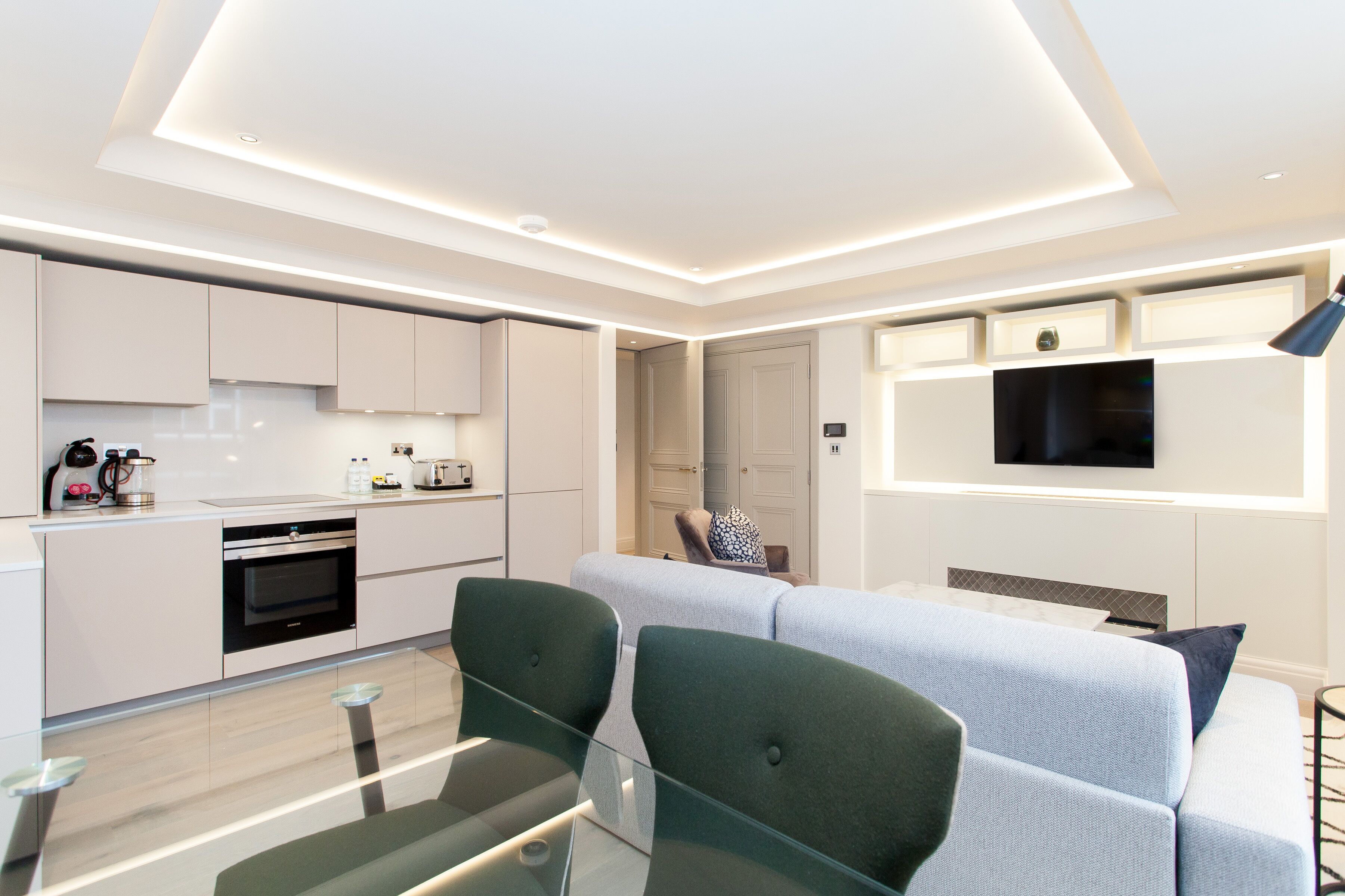 A modern kitchen with built-in appliances, a dining area with a glass table, and a living area with a sofa and TV.