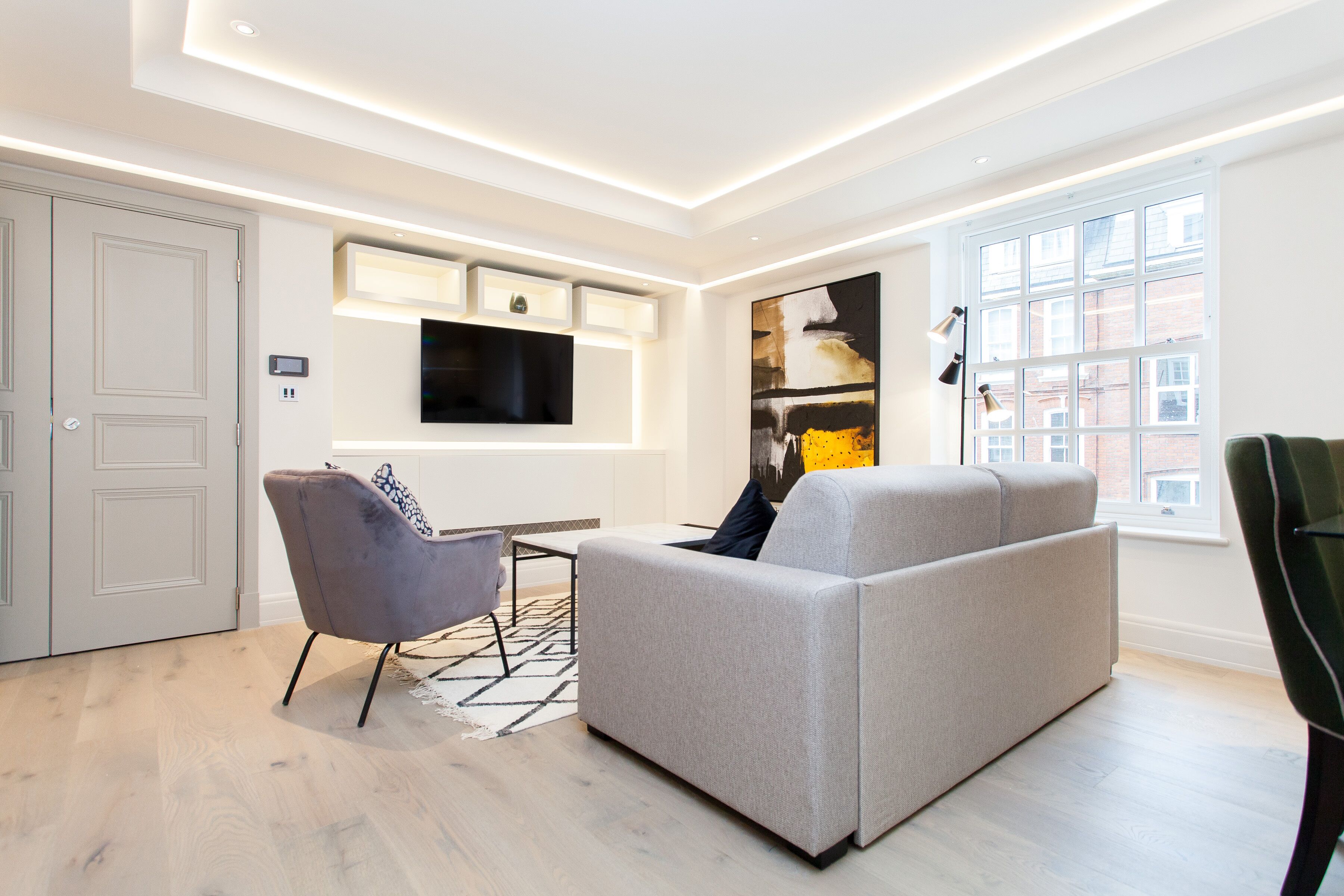 A modern living room with a grey sofa, a grey armchair, and a flat-screen TV mounted on the wall. There is a window with a view of buildings outside.