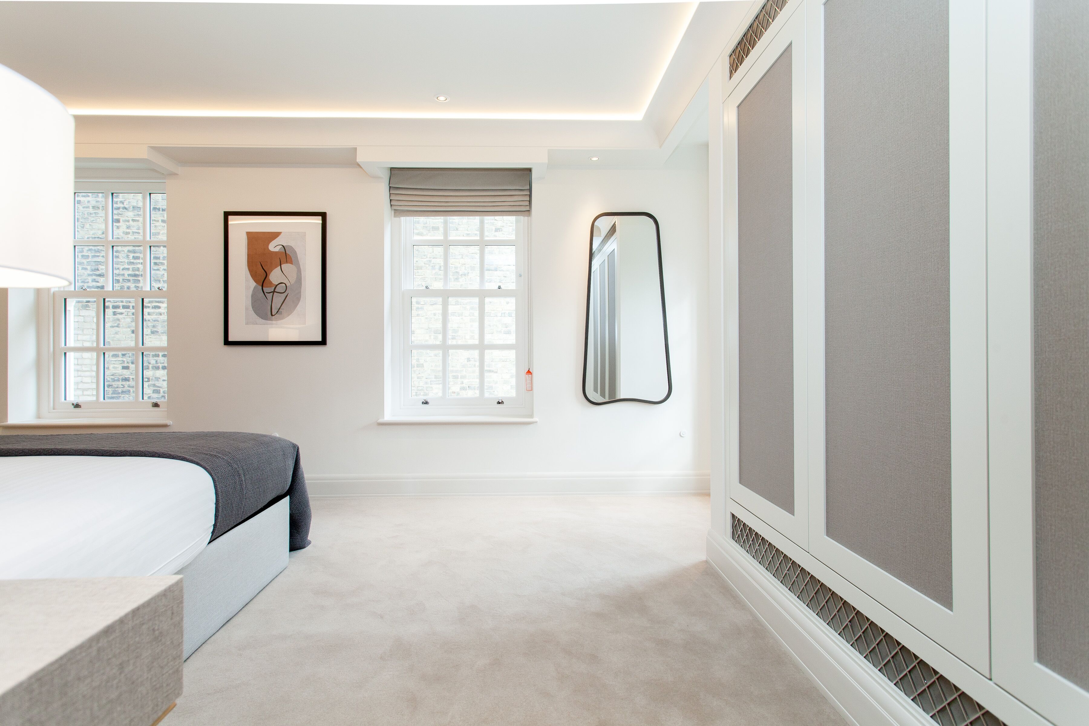 A modern bedroom with a large bed, a bedside table, a window with blinds, a framed artwork, and a large mirror.