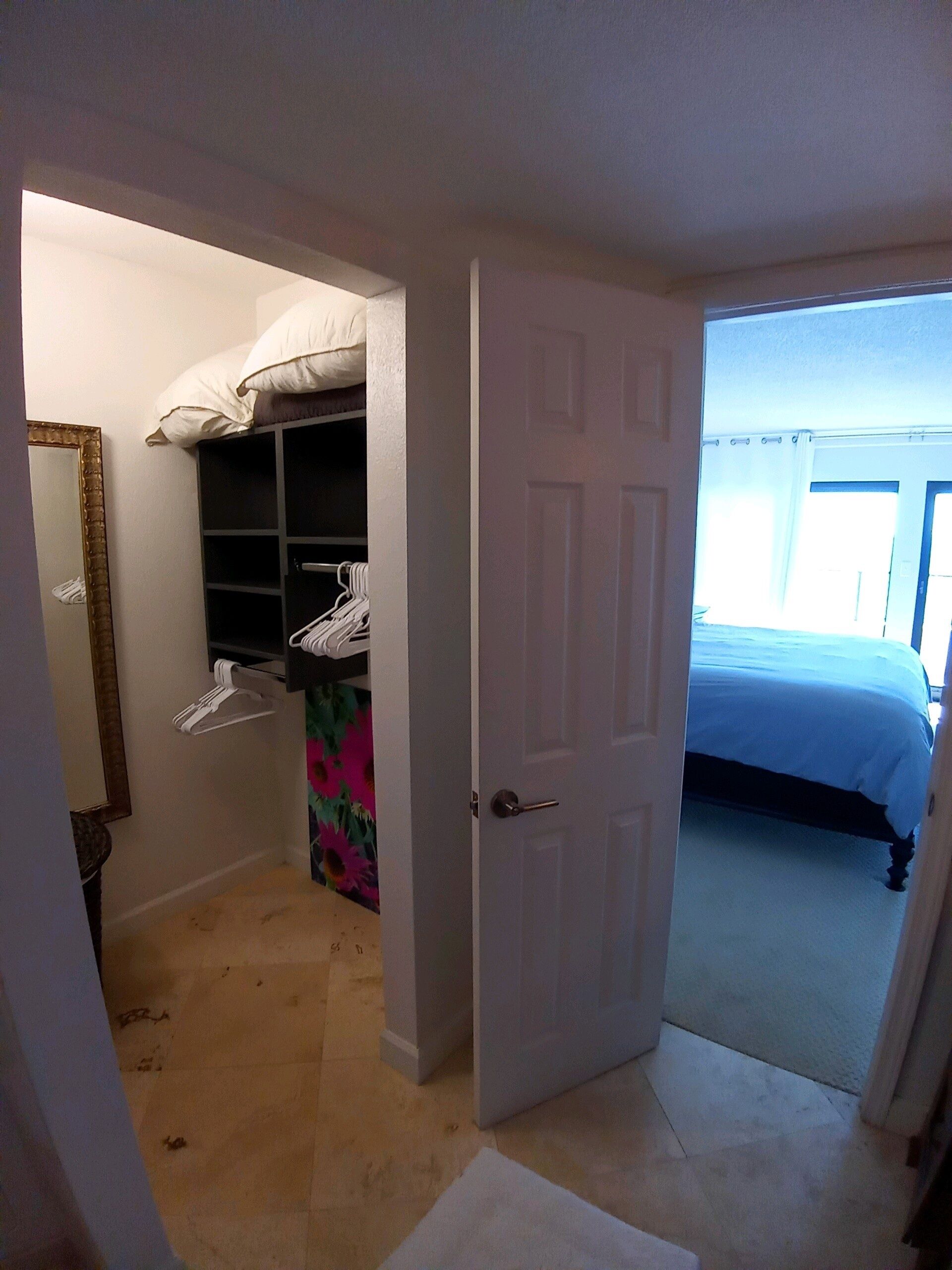 Une chambre à coucher avec un lit, une armoire où sont suspendus des vêtements, et une porte qui donne sur une autre pièce.