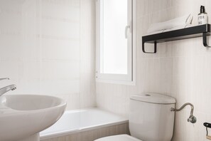 A bathroom with a white sink, toilet, and bathtub. There is a window providing natural light and a shelf with toiletries.