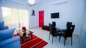 A modern living room with a glass coffee table, blue sofa, red and black striped rug, and a dining area with black chairs and a red door. - Complexe Hôtelier Akparo (Dabou)