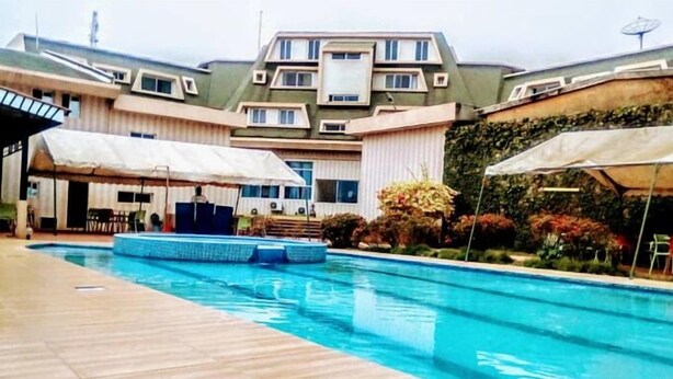 A swimming pool with a wooden deck, umbrellas, and a building with a green roof and multiple windows. - Complexe Hôtelier Akparo (Dabou)