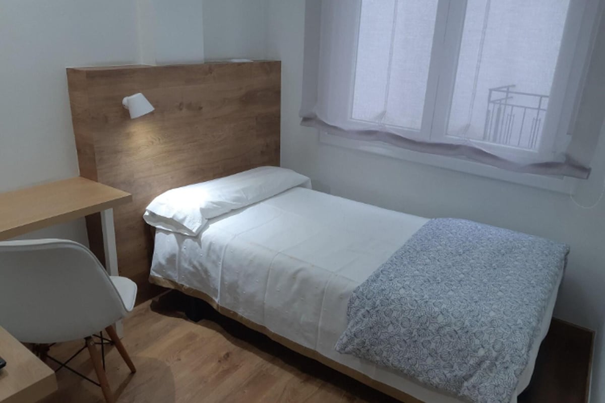 Una cama individual con cabecera de madera, un escritorio pequeño con una silla, y una ventana con vista a un balcón.