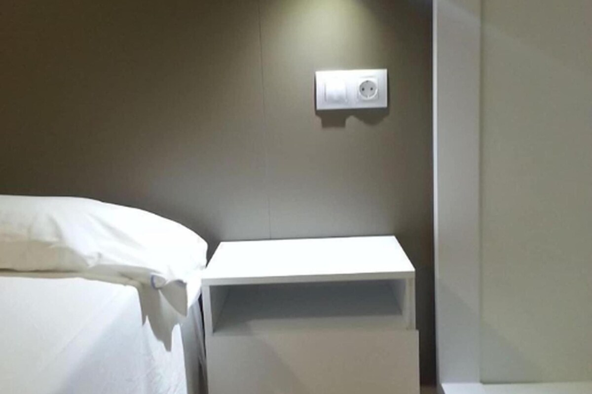 Un dormitorio pequeño y ordenado con una cama individual, una mesita de noche con lámpara y un enchufe en la pared.