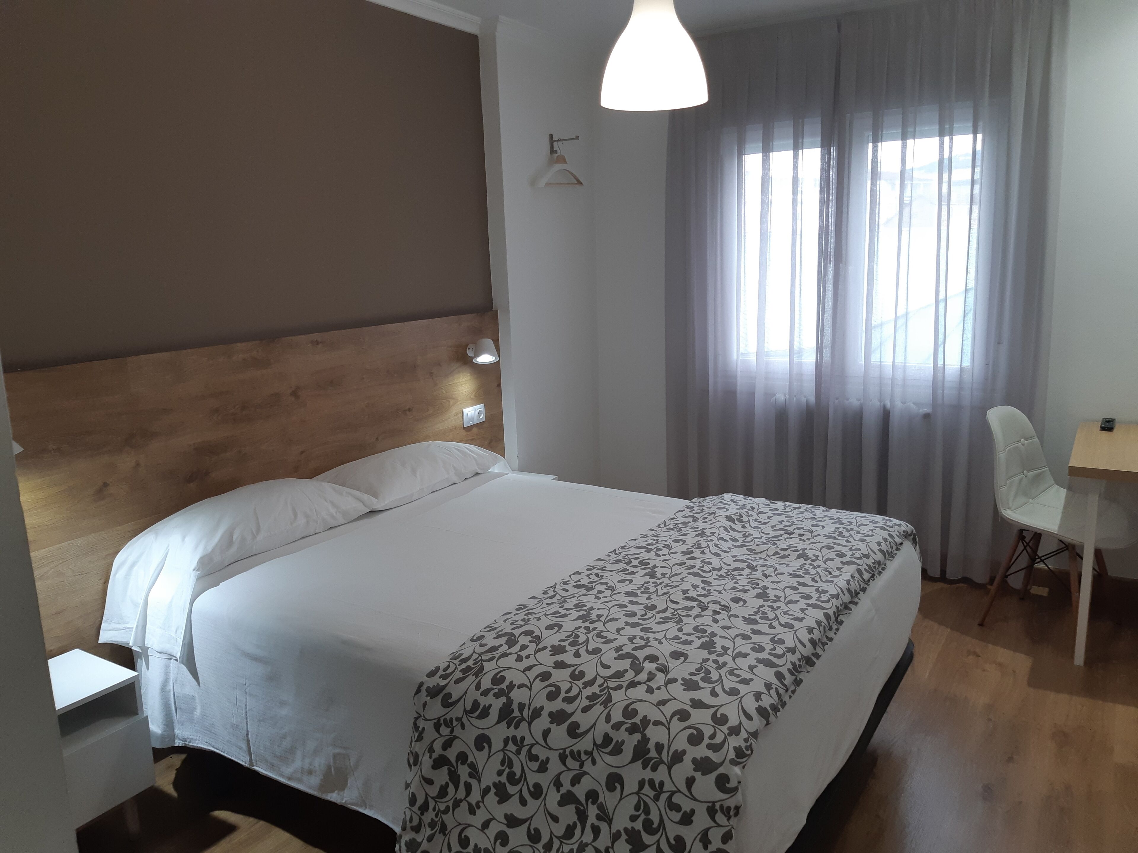 A neatly made bed with a patterned bedspread, a wooden headboard, a bedside table, a chair, and a window with sheer curtains.