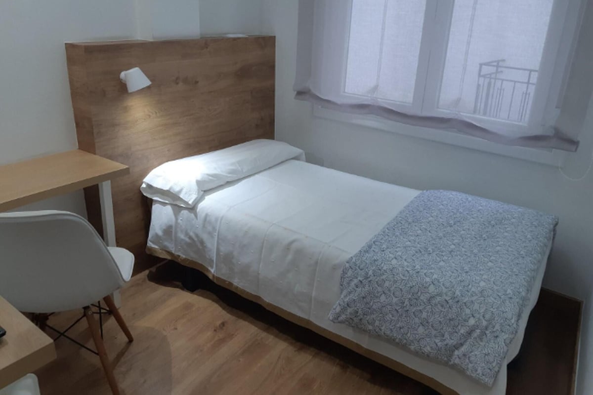 Ein Einzelbett mit Holz Kopfteil, ein kleiner Schreibtisch mit Stuhl und ein Fenster mit Blick auf einen Balkon.