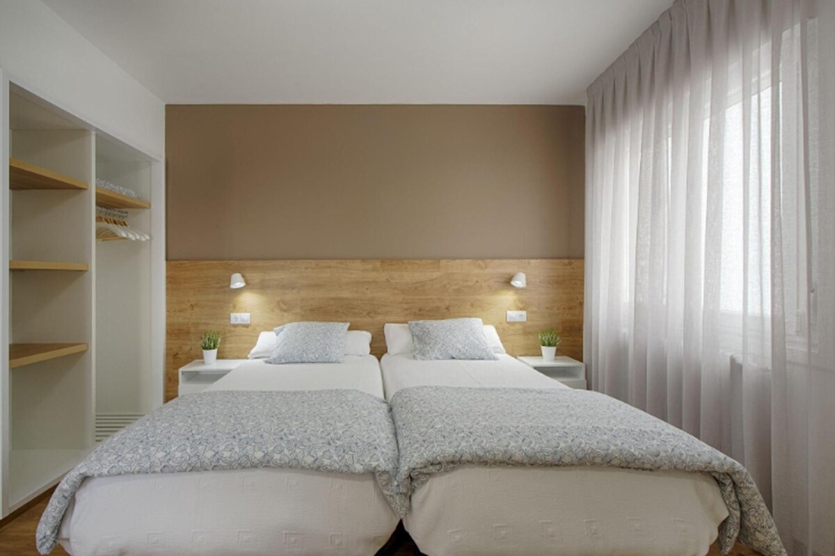 A hotel room with two beds, a wooden headboard, bedside tables with lamps, a wardrobe, and sheer curtains.