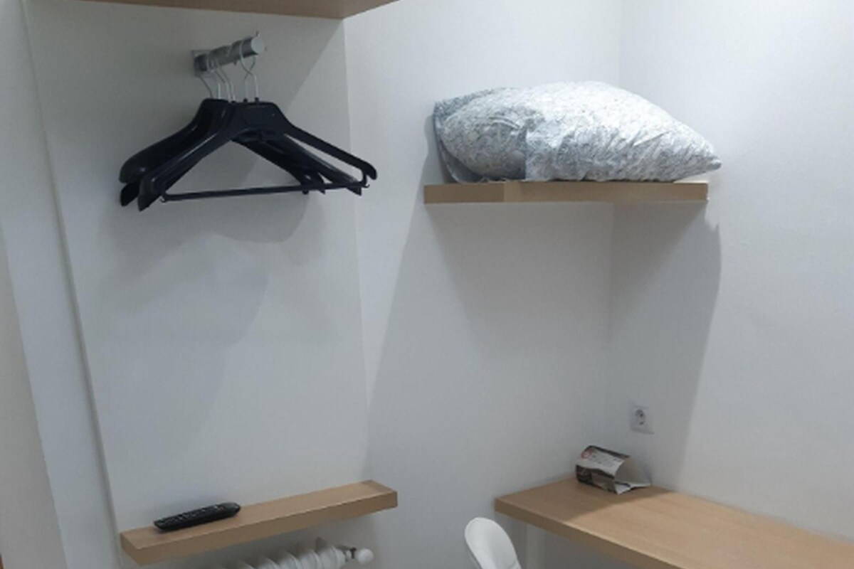 A small, neatly organized room with a wooden desk, a white chair, a radiator, a hanging black hanger, and a pillow on a shelf.