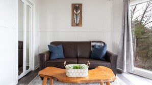 Un salon avec un canapé en cuir marron, deux coussins bleus, une table basse en bois surmontée d’un pot de fleurs blanc, et une grande fenêtre avec des rideaux.