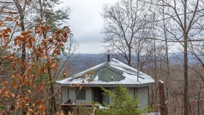 Une petite cabane à un seul étage, entourée d’arbres, avec une terrasse en bois et une clôture en métal.