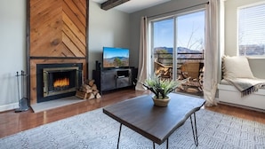 Ein Wohnzimmer mit Kamin, Fernseher, Esstisch und einem großen Fenster mit Bergblick.