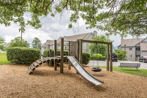 A playground with a slide and swings, surrounded by a wooden fence and mulch, with houses in the background. - Unreal Mt. Leconte Views/Indoor Pool And Hot Tub (Gatlinburg)