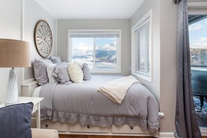 Une chambre à coucher avec un lit, une fenêtre avec des stores, un élément décoratif fixé au mur et une vue sur le ciel.