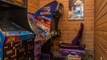 A small room with wooden walls and a tiled floor, containing a vintage arcade machine, a purple gaming console, and a Nintendo Wii console.