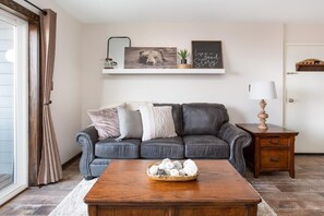A living room with a dark leather sofa, a wooden coffee table, and a small wooden nightstand with a lamp.