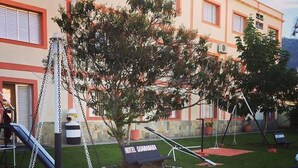 Un parque infantil con tobogán y columpio, rodeado de un césped cuidado y un edificio con un letrero que dice 'HOTEL GUARANAHMA'.