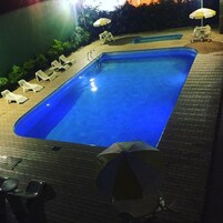 Piscina retangular com esguicho e deck ladrilhado, rodeada por espreguiçadeiras brancas e guarda-sóis.