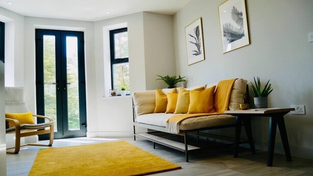 A modern living room with a sofa, yellow cushions, a coffee table, and a yellow rug. There are two windows with curtains, a door, and framed artwork on the wall. - ⭐⭐⭐⭐⭐ Beautiful Apartment Near the BEACH +Netflix (Bournemouth)