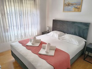 A neatly made bed with white linens and a pink accent, a headboard, a nightstand with a lamp, and a painting of a coastal scene on the wall. - Apartments Pepa (Rogoznica)