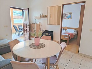 A dining area with a round table, chairs, a vase with flowers, a television, and a bed in the adjacent room. - Apartments Pepa (Rogoznica)