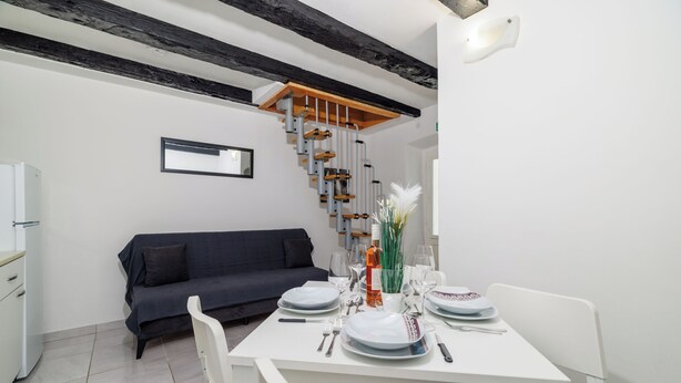 A dining area with a white table set for four, a dark sofa, and a wooden staircase with exposed beams. - Apartment Lozann (Povile)