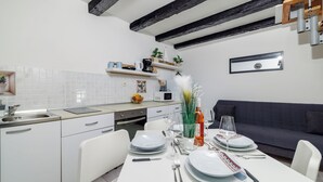 A modern kitchen with white cabinets, a dining table set for four, and a dark blue sofa in the adjacent living area. - Apartment Lozann (Povile)