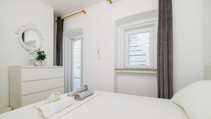 A neatly made bed with rolled towels, a white dresser, a mirror, and a window with curtains. - Apartment Lozann (Povile)