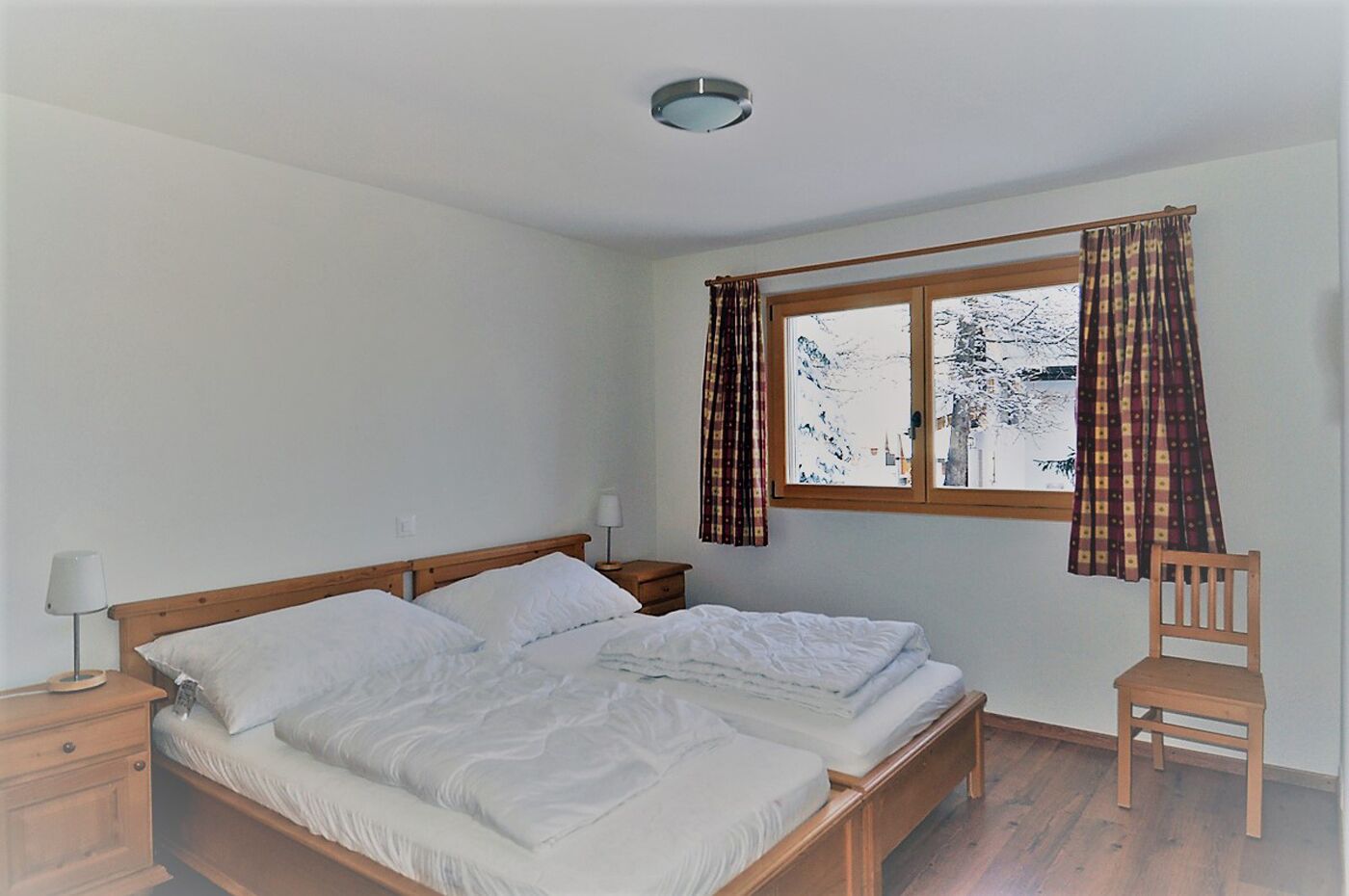 A bedroom with a wooden bed, a nightstand, a chair, and a window with curtains.