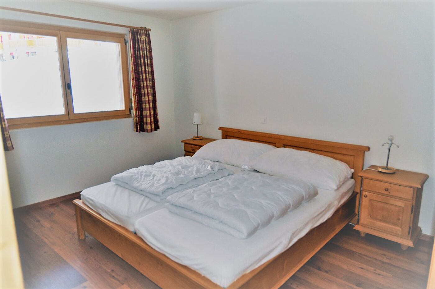 A bedroom with a wooden bed, two bedside tables, a window with curtains, and a wooden floor.