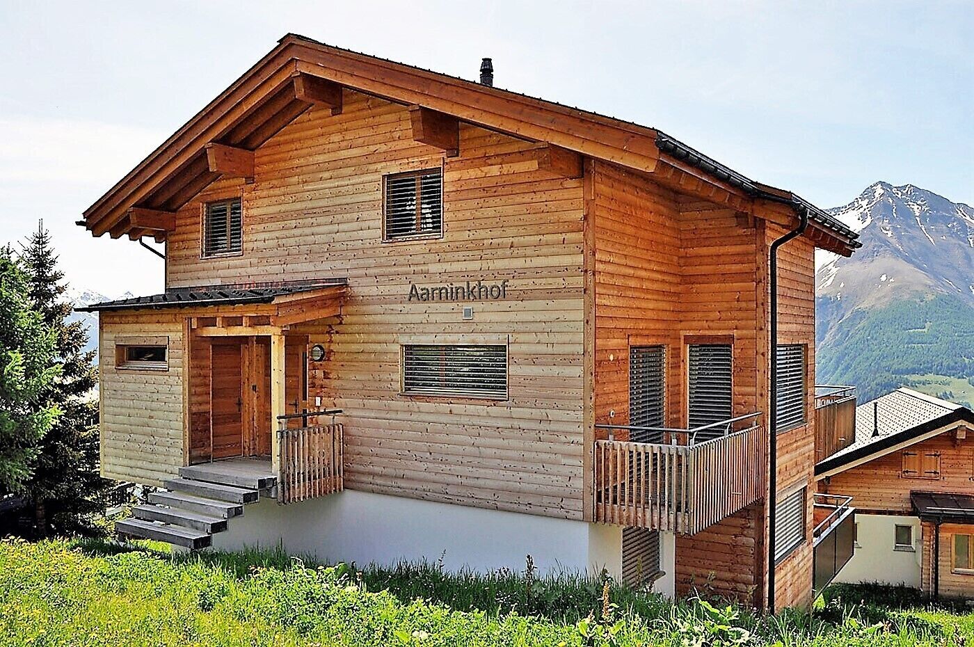 A wooden chalet with a balcony, a staircase leading to the entrance, and a mountain view in the background.