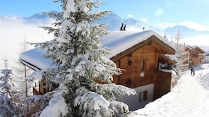 Um chalé de madeira com o telhado coberto de neve, rodeado por árvores e montanhas também cobertas de neve ao fundo.