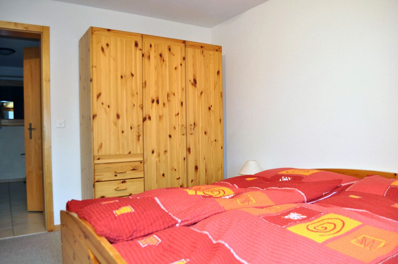 A bedroom with a wooden wardrobe, a bed with a red patterned bedspread, and a white wall.