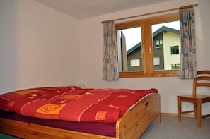 A bedroom with a wooden bed, a window with curtains, and a chair. - Alpengarten 4 (Bettmeralp)
