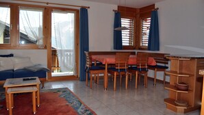 Una sala de estar acogedora con un sofá, una mesa de comedor con sillas, una pequeña cocina y ventanas amplias con cortinas.