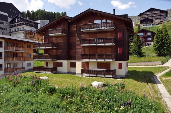 Un edificio de madera de varios pisos con balcones, rodeado de vegetación y otros edificios.