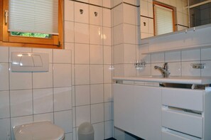 A modern bathroom with white tiles, a toilet, a sink with a mirror, and a window with blinds.