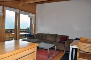A cozy living room with a wooden table, a sofa, and a view of mountains through large windows.