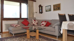 Salón con sofá gris, cojines estampados en rojo y blanco, mesa de centro de vidrio, radiador, una silla de madera y dos cuadros en la pared.