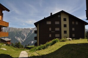 A two-story building with a dark facade and a sign reading "Alpenglück." It is situated on a grassy slope with a mountain view in the background. - Alpenglück 43 (Bettmeralp)
