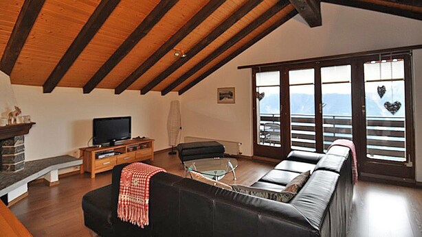 A living room with a wooden ceiling, a black sofa, a glass coffee table, and a fireplace. - Alpenglück 51 (Bettmeralp)