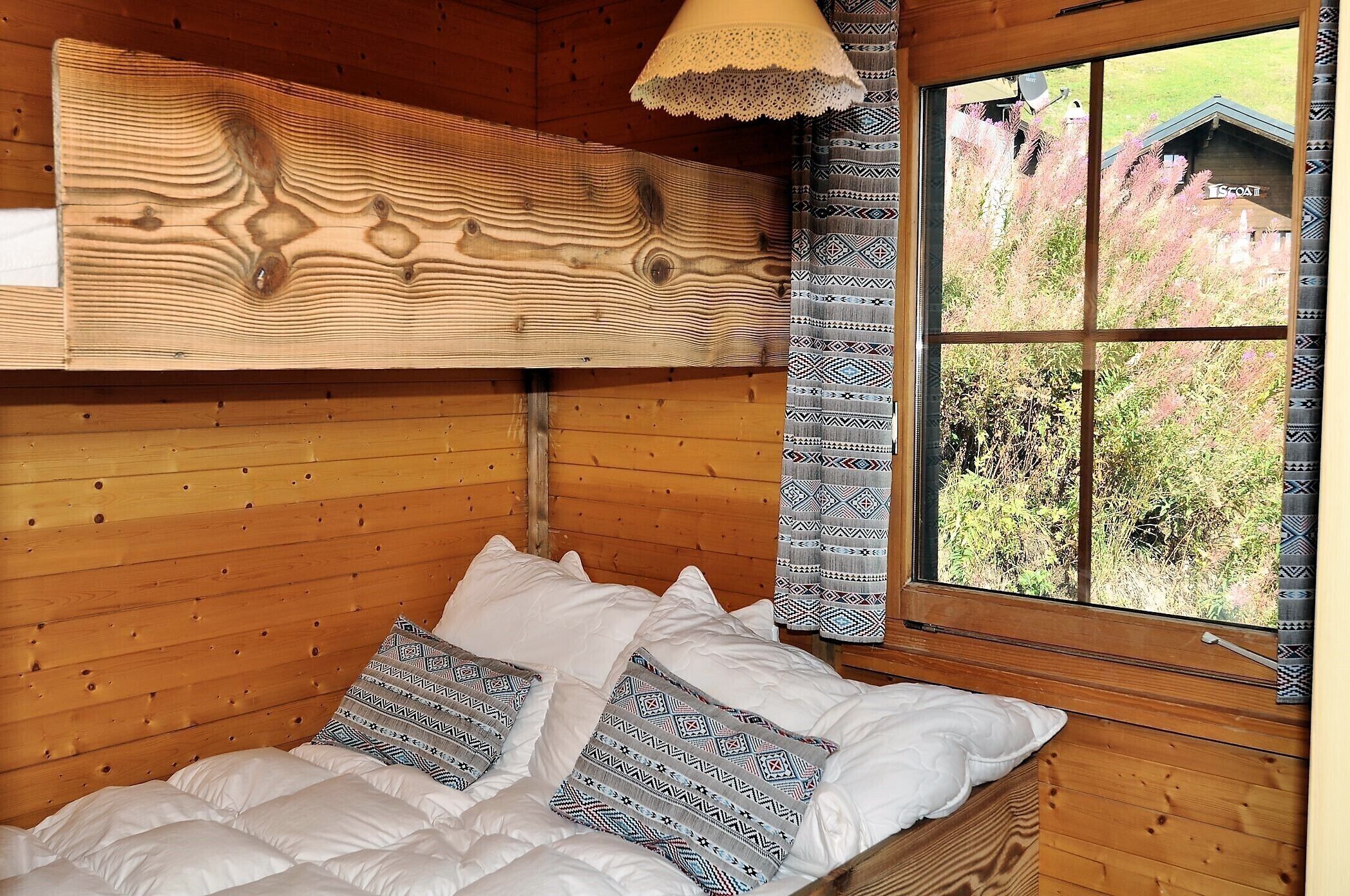Une pièce de style cabane en bois, avec un lit, des coussins à motifs, une fenêtre donnant sur la verdure et un plafond orné de panneaux de bois.