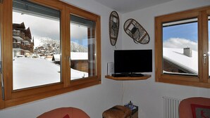 Una stanza con una TV su uno scaffale, finestre con cornice in legno e vista su un villaggio montano innevato.
