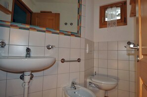 A bathroom with a white sink, a toilet, and a bidet. There is a mirror above the sink and a window with a wooden frame.