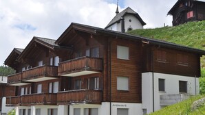 A multi-story wooden building with balconies, a church with a steeple, and a sign reading "Ambiente-B". - Ambiente B21 (Bettmeralp)