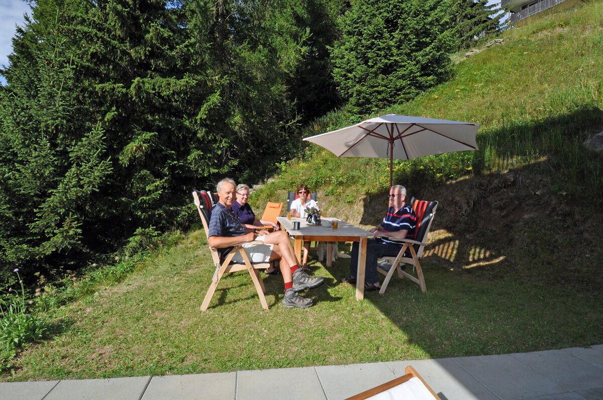 A group of people seated at an outdoor table with an umbrella, surrounded by greenery.