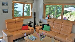 A living room with two leather sofas, a coffee table with magazines and a vase of flowers, and large windows with a view of trees and mountains. - Bettmerlodge (Bettmeralp)