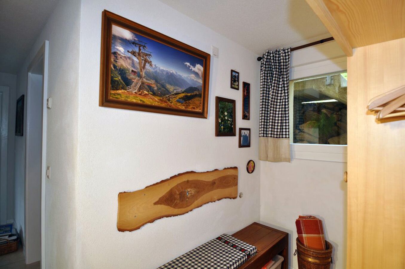 Una habitación con un cuadro grande de un paisaje enmarco, un adorno de pared de madera, una pequeña mesa con mantel a cuadros y una ventana con cortinas a cuadros.