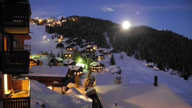 Un pueblo cubierto de nieve con edificios iluminados, una luna destacada en el cielo y una colina boscosa al fondo.