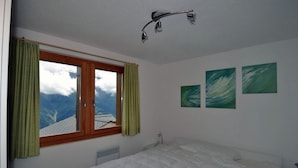 A bedroom with a bed, a window with curtains, a radiator, and three green abstract paintings on the wall. - Camelot 4 (Bettmeralp)